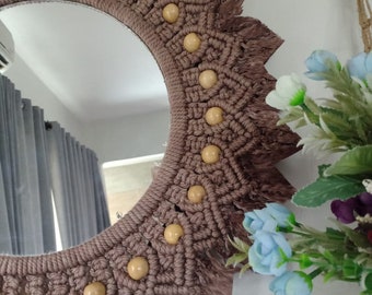 Peut inclure: Un miroir en macramé marron avec un cadre circulaire et des perles en bois. Le miroir a une bordure en frange et est suspendu à un mur blanc.
