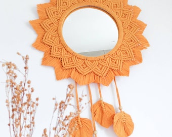 Peut inclure: Un miroir rond avec un cadre en macramé de couleur orange vif. Le cadre est décoré de glands et de plumes.