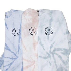 Op de afbeelding: Drie tie-dye sweatshirts in blauw, roze en lichtblauw met een zwart smiley-gezicht en het woord "smile" eromheen herhaald.