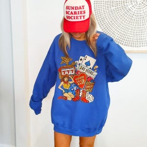 May include: A royal blue sweatshirt with a Las Vegas-themed graphic, including playing cards, dice, and a cowgirl. The sweatshirt is paired with brown cowboy boots and a red baseball cap with the text "SUNDAY SCARIES SOCIETY".