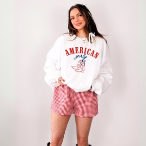 Puede incluir: Sudadera blanca con texto rojo que dice "AMERICAN girly" y un gráfico de botas de vaquero rojas. La modelo lleva pantalones cortos de cuadros rojos y blancos.