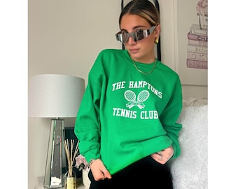May include: A green sweatshirt with white text that reads "The Hamptons Tennis Club" and a graphic of two tennis rackets. The sweatshirt is worn by a person wearing black pants and sunglasses.
