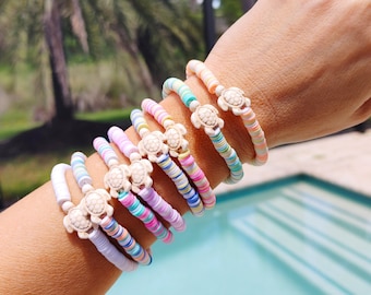 Puede incluir: Una pila de pulseras coloridas con un dije de tortuga blanca en cada una. Las pulseras están hechas de pequeñas cuentas redondas y están superpuestas en la muñeca de una persona.