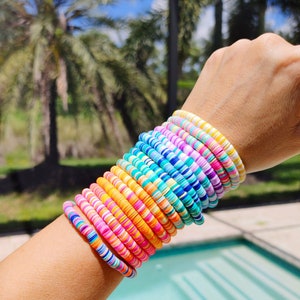 Op de afbeelding: Een stapel kleurrijke armbanden gemaakt van kleine, ronde kralen. De armbanden zijn gelaagd in een regenboogpatroon, met tinten roze, oranje, geel, blauw, groen en wit.