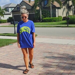 May include: A woman wearing a blue oversized t-shirt with the text "SUNKISSED" in yellow. She is wearing brown sandals and walking on a brick pathway.