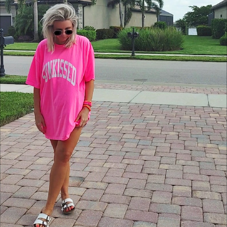 Puede incluir: Una mujer lleva una camiseta rosa de gran tama&ntilde;o con la palabra "SUNKISSED" impresa en blanco en la parte delantera. Lleva sandalias blancas y gafas de sol.