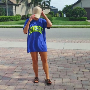 Puede incluir: Una persona lleva una camiseta azul con la palabra "SUNKISSED" en letras amarillas. Lleva una gorra de b&eacute;isbol beige y sandalias marrones.