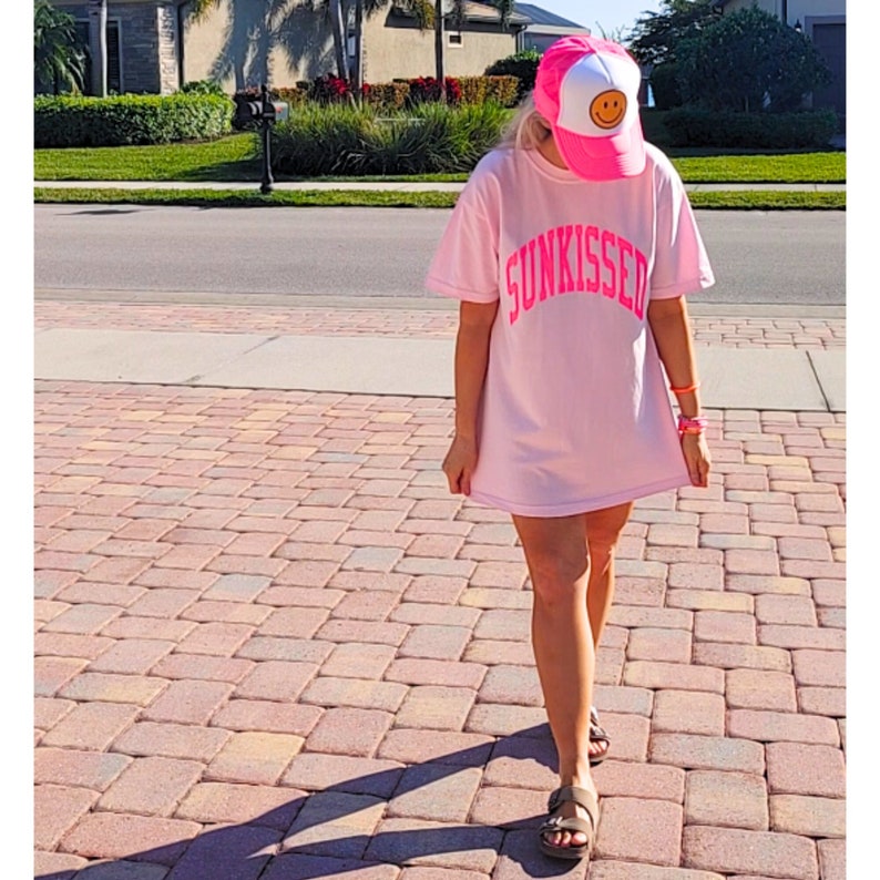 May include: A pink oversized t-shirt with the word "SUNKISSED" printed in neon pink. The person is wearing a pink and white trucker hat with a yellow smiley face.
