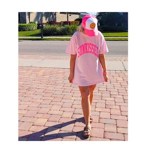 May include: A pink oversized t-shirt with the word "SUNKISSED" printed in pink on the front. The person is wearing a pink and white baseball cap with a yellow smiley face on it.