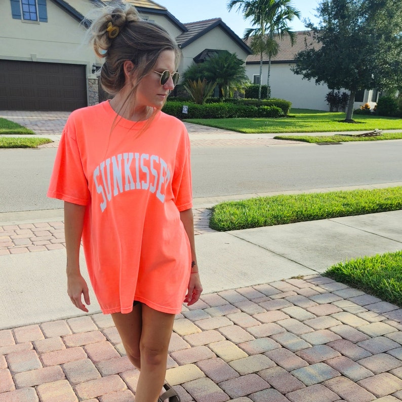 May include: A woman wearing a bright pink t-shirt with the word "Sunkissed" printed in white on the front. She is standing on a brick walkway.