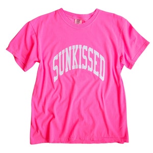 May include: A bright pink t-shirt with the word "SUNKISSED" printed in white in an arch across the front.