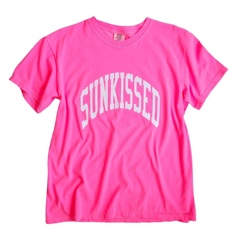 May include: A bright pink t-shirt with the word "SUNKISSED" printed in white in an arch across the front.