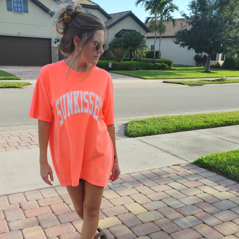 Puede incluir: Una mujer lleva una camiseta naranja brillante con el texto "SUNKISSED" impreso en blanco en la parte delantera. Est&aacute; de pie en un camino de adoquines.