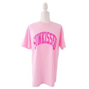 May include: Pink t-shirt with a pink "SUNKISSED" graphic in an arch across the front.