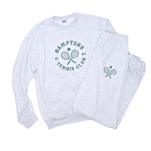 May include: Gray sweatshirt and sweatpants set with a green tennis racket and ball graphic. The sweatshirt has the text "HAMPTONS TENNIS CLUB 19 83" around the graphic. The sweatpants have a smaller version of the graphic on the left leg.