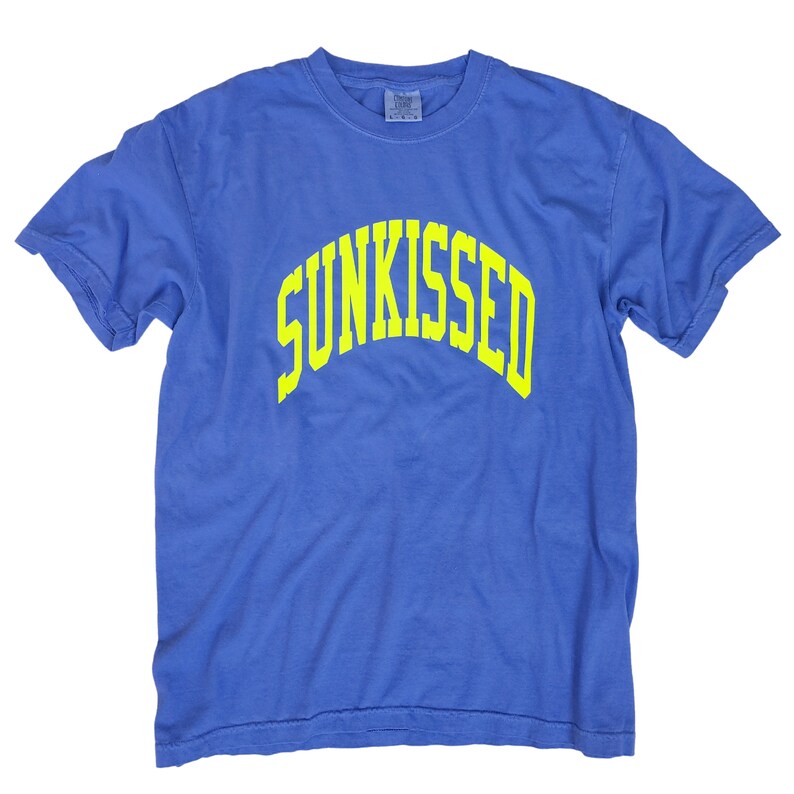 Puede incluir: Una camiseta azul con un estampado amarillo "SUNKISSED" en la parte delantera.