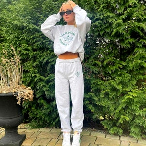 May include: A gray sweatshirt with the text "Champion Tennis Club" and a graphic of two tennis rackets. The sweatshirt is paired with gray sweatpants with a small graphic of two tennis rackets on the left leg.