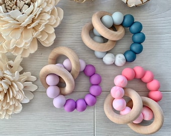 May include: Three wooden teething rings with silicone beads. The rings are natural wood and the beads are in shades of purple, pink, and blue.
