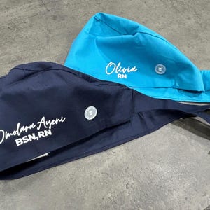 May include: Two scrub caps, one navy blue and one turquoise. The navy blue cap has the name "Omolara Ayeni" and "BSN, RN" printed in white. The turquoise cap has the name "Olivia" and "RN" printed in white. Both caps have a button closure.