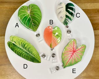 Puede incluir: Colección de cinco portatarjetas con temática botánica. Los portatarjetas presentan diseños de hojas en varias formas y colores, incluyendo verde, rosa y blanco. Cada portatarjetas tiene un cordón retráctil de plástico transparente y un clip metálico.