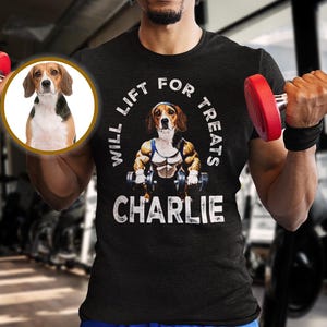 Może przedstawiać: Czarny t-shirt z napisem "WILL LIFT FOR TREATS CHARLIE" i kreskówkowym beagle podnoszącym ciężary. Osoba na zdjęciu ma na sobie niebieskie szorty i podnosi czerwone hantle. Zdjęcie beagle'a znajduje się w okręgu.