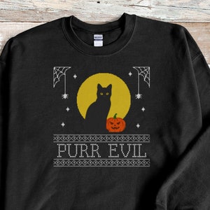 May include: A black crewneck sweatshirt featuring a graphic print of a black cat sitting in front of a yellow full moon next to an orange jack-o'-lantern. The design includes white spiderwebs, small stars, and the text PURR EVIL in white letters.