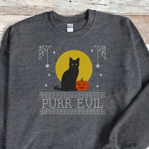 May include: A dark heather gray crewneck sweatshirt featuring a graphic print of a black cat sitting next to an orange jack-o'-lantern in front of a yellow moon. The design includes white spiderwebs and the text PURR EVIL in a block font.