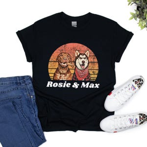Graphic t-shirt featuring a pitbull and husky portrait with the names in bold, white, distressed font below. The dog image is placed inside a vintage-style, sunset-colored circle, blending shades of orange, yellow, and red for a retro aesthetic.