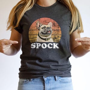 May include: Dark gray t-shirt featuring a graphic of a French Bulldog with the name "SPOCK" in white text. The dog illustration is set against a sunset-style background with orange, yellow, and brown hues.
