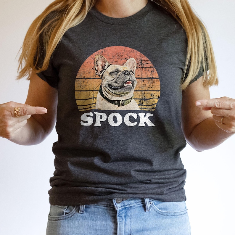 May include: Dark gray t-shirt featuring a graphic of a French Bulldog with the name "SPOCK" in white text. The dog illustration is set against a sunset-style background with orange, yellow, and brown hues.