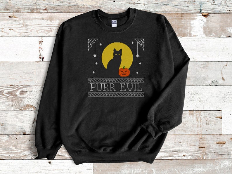 May include: A black crewneck sweatshirt featuring a graphic print of a black cat sitting in front of a yellow full moon next to an orange jack-o'-lantern. The design includes white spiderwebs, small stars, and the text PURR EVIL in white letters.