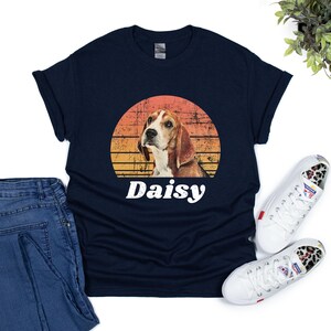 Graphic t-shirt featuring a beagle dog portrait with the name in bold, white, distressed font below. The dog image is placed inside a vintage-style, sunset-colored circle, blending shades of orange, yellow, and red for a retro aesthetic.