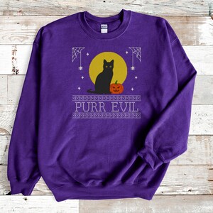May include: A purple long-sleeve sweatshirt featuring a graphic print of a black cat sitting in front of a yellow moon next to an orange jack-o'-lantern. The design includes white spiderwebs and the text PURR EVIL in a decorative border.