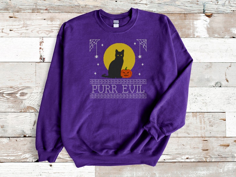 May include: A purple long-sleeve sweatshirt featuring a graphic print of a black cat sitting in front of a yellow moon next to an orange jack-o'-lantern. The design includes white spiderwebs and the text PURR EVIL in a decorative border.