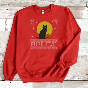May include: A red crewneck sweatshirt featuring a graphic print of a black cat sitting next to an orange jack-o'-lantern in front of a yellow moon. The design includes white spiderwebs, stars, and the text PURR EVIL in a decorative border.