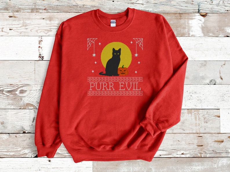 May include: A red crewneck sweatshirt featuring a graphic print of a black cat sitting next to an orange jack-o'-lantern in front of a yellow moon. The design includes white spiderwebs, stars, and the text PURR EVIL in a decorative border.