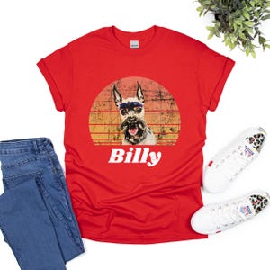 Graphic t-shirt featuring a dog portrait with the name in bold, white, distressed font below. The dog image is placed inside a vintage-style, sunset-colored circle, blending shades of orange, yellow, and red for a retro aesthetic.