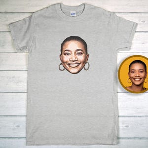 May include: A pale grey t-shirt with a portrait of a smiling person wearing hoop earrings. A smaller, circular image of the same portrait is on the right. The shirt is made of a soft material.