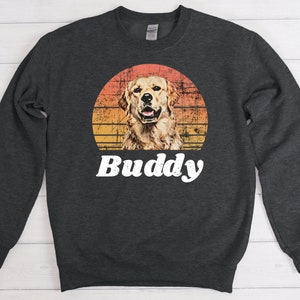 May include: A dark gray sweatshirt with a retro sunset graphic featuring a golden retriever dog. The text "Buddy" is printed in white below the graphic.