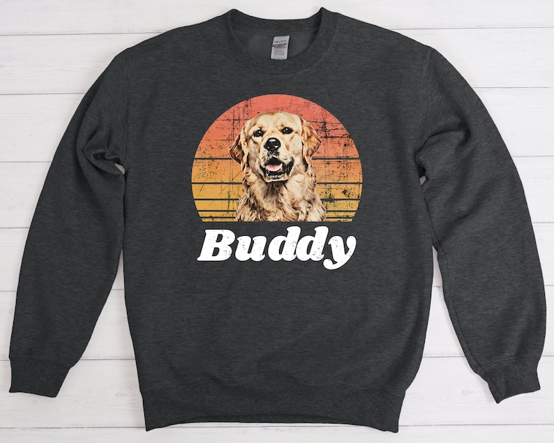 May include: A dark gray sweatshirt with a retro sunset graphic featuring a golden retriever dog. The text "Buddy" is printed in white below the graphic.