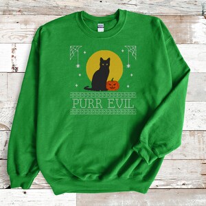 May include: A bright green crewneck sweatshirt featuring a graphic print of a black cat sitting next to an orange jack-o'-lantern in front of a yellow moon. The design includes white spiderwebs and the text PURR EVIL in a stylized font.