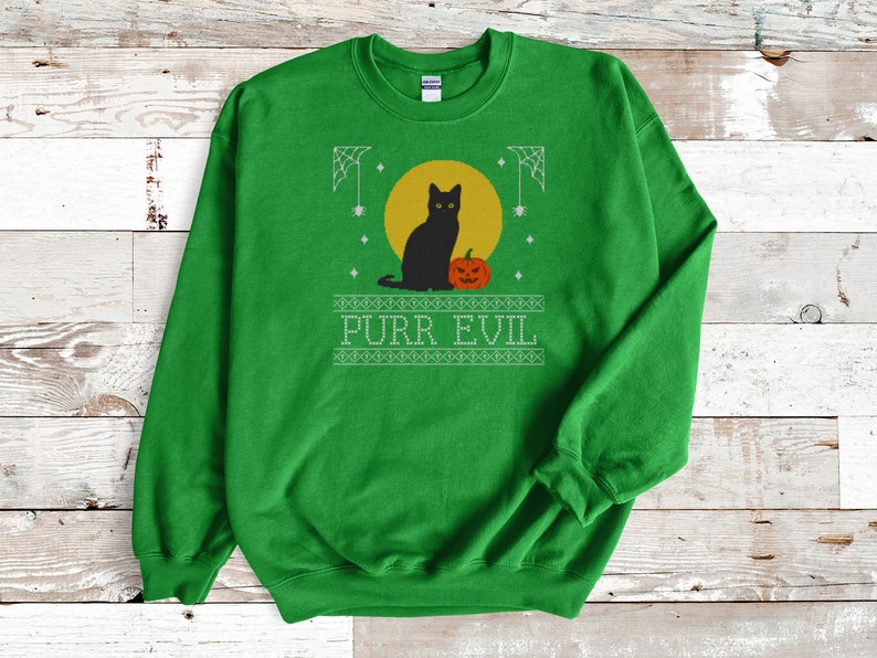 May include: A bright green crewneck sweatshirt featuring a graphic print of a black cat sitting next to an orange jack-o'-lantern in front of a yellow moon. The design includes white spiderwebs and the text PURR EVIL in a stylized font.