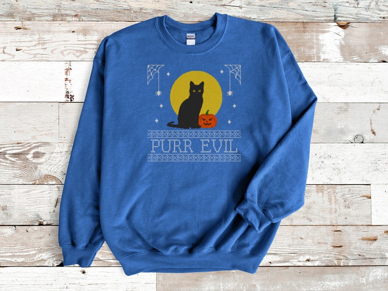 May include: A royal blue crewneck sweatshirt featuring a graphic print of a black cat sitting next to an orange jack-o'-lantern in front of a yellow full moon. The design includes white spiderwebs, stars, and the text PURR EVIL in a block font.