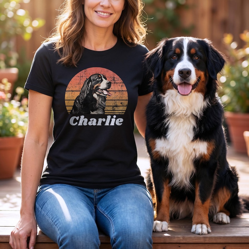 Black personalized dog photo T-shirt mockup showing a smiling woman in jeans sitting outdoors beside a Bernese Mountain Dog, with a retro sunset portrait design and the name Charlie on the shirt.