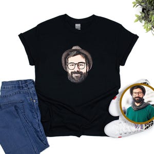May include: Black t-shirt with a portrait of a man wearing glasses, a hat, and a beard. The image is paired with blue jeans and white trainers. A circular image of the man is also present, showing a green scarf.