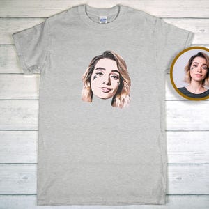 May include: A pale grey t-shirt with a portrait of a woman with blonde hair and a star-shaped facial marking. A smaller, circular image of the same woman is visible on the right. The t-shirt is made of a soft material.