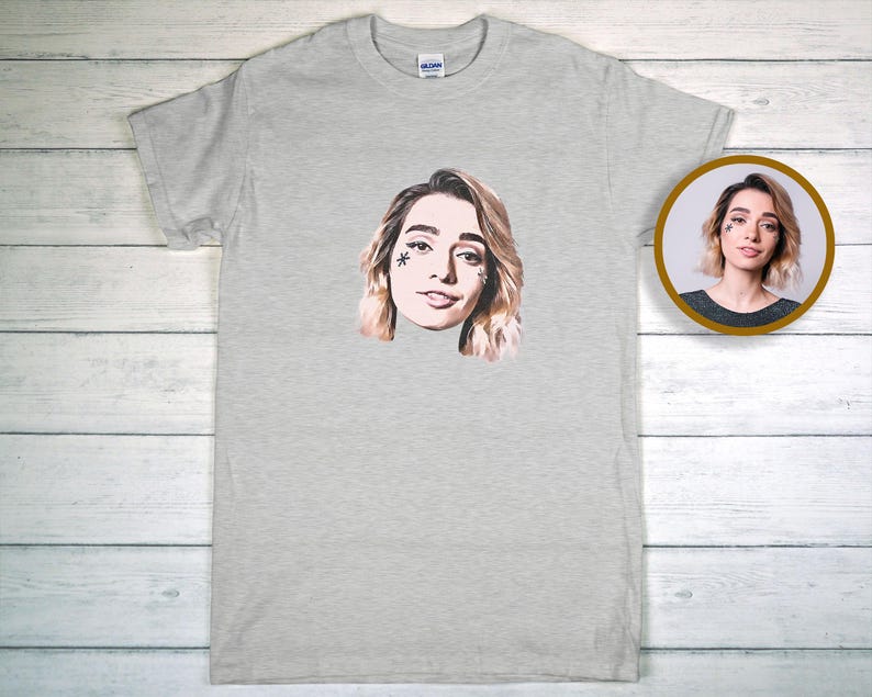 May include: A pale grey t-shirt with a portrait of a woman with blonde hair and a star-shaped facial marking. A smaller, circular image of the same woman is visible on the right. The t-shirt is made of a soft material.