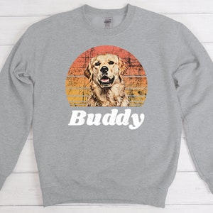 May include: A gray sweatshirt with a retro sunset graphic featuring a golden retriever dog. The text "Buddy" is printed below the graphic.