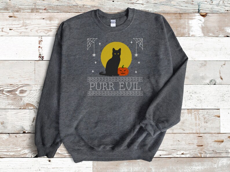 May include: A dark heather gray crewneck sweatshirt featuring a graphic print of a black cat sitting next to an orange jack-o'-lantern in front of a yellow moon. The design includes white spiderwebs and the text PURR EVIL in a block font.