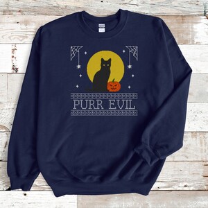 May include: A navy blue crewneck sweatshirt featuring a graphic print of a black cat sitting in front of a yellow full moon next to an orange jack-o'-lantern. The design includes white spiderwebs, stars, and the text PURR EVIL in a block font.
